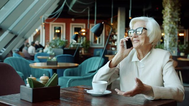 Happy senior woman wearing glasses is discussing business on mobile phone working at table in cafe. Communication and elderly people concept.