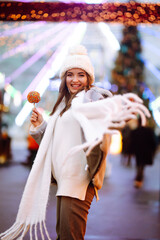 Obraz premium Young woman with caramel apple on Christmas market. Smiling woman in winter style clothes posing at festive street market. Lights around. Winter holiday.