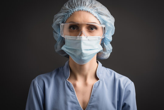 nurse in medical cap, mask and goggles looking at camera isolated on dark grey, stock image
