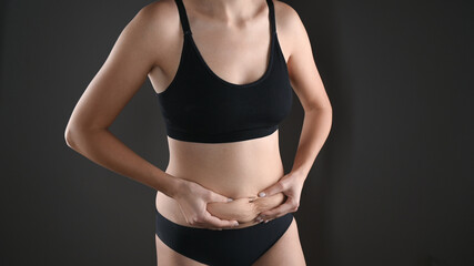 Fototapeta premium Woman in black underwear holding her fat belly, stomach with hands, diet, weight loss and liposuction concept. Isolated on grey background