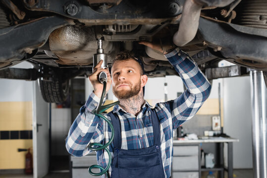 mechanic fixing detail with pneumatic wrench while standing under raised car, stock image