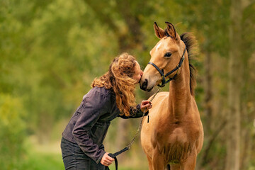 A light brown buckskin foal, woman lovingly kisses the foal on his nose. autumn colors