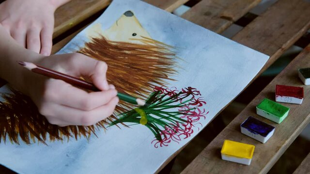 little girl draws a hedgehog with flowers on the back on a sheet of paper with watercolors and pencils, children's creativity and drawing, concept