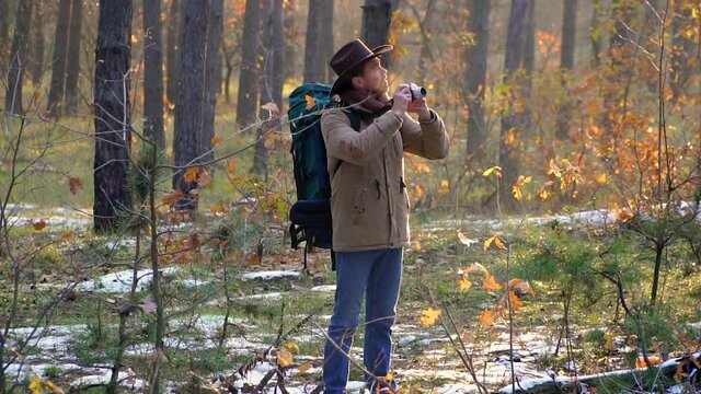 A man in a hat with a backpack takes pictures in the forest. Beautiful autumn forest and the first snow. Travel concept. Slow motion