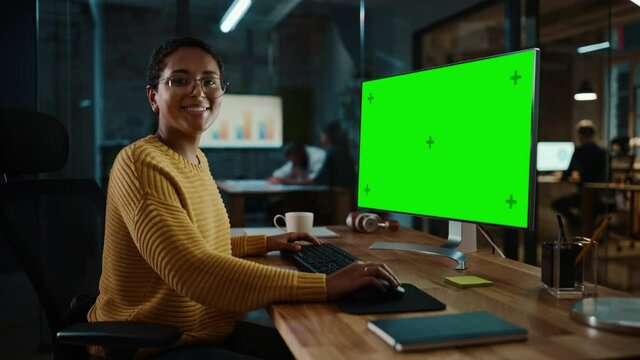 Young Multiethnic Specialist Working on Desktop Computer with Green Screen Mock Up Display in Creative Office. Beautiful Diverse Female Manager with Short Hair and Glasses is Wearing a Yellow Jumper.