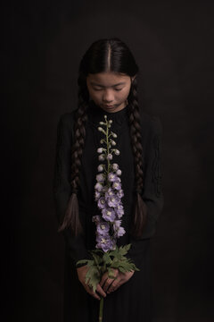 Classic painterly portrait of a girl with black braids holding a purple larkspur (delphinium) flower in her hands in dark studio setting