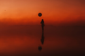 surreal silhouette of a girl with a balloon in her hand on the water at sunrise in the fog in summer