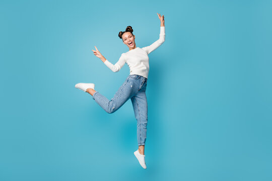 Full length photo of girl jump laugh show two v-signs wear white pullover jeans sneakers isolated blue color background