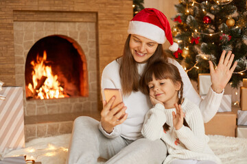 Mom and daughter sitting near Christmas tree, hugging, communicating via video call, having fun. Christmas, new year's eve at home. Little girl with mother near fireplace.