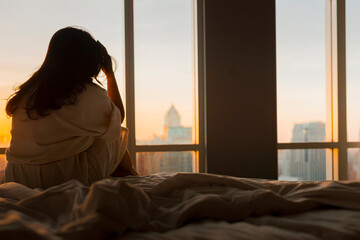 Unhappy woman touching hair, sitting on the bed beside the windows with sunlight in the morning, thinking about problems, upset girl feeling lonely and sad, suffering from bad relationship or break up