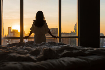 Beautiful asian woman is waking up in the morning, standing near bed, Sun shines on her from the big window. Happy young girl greets new day with warm sunlight flare and city scenery in the window.