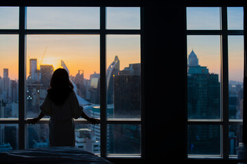 Fototapeta na wymiar Young happy woman standing in the bedroom by the window with her back and beautiful sunrise or sunset and city scenery in the window. Copy space