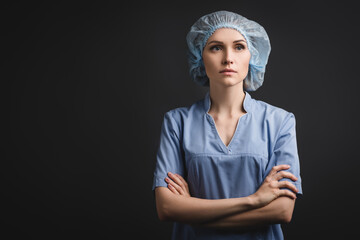 nurse in medical cap standing with crossed arms and looking away isolated on dark grey, stock image
