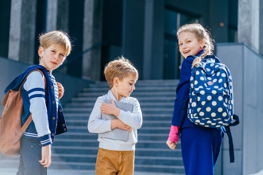 Back view of three schoolchildren standing near their school. Two boys and girl in uniform and backpacks. Pupils with books and backpacks outdoors with school building on background.