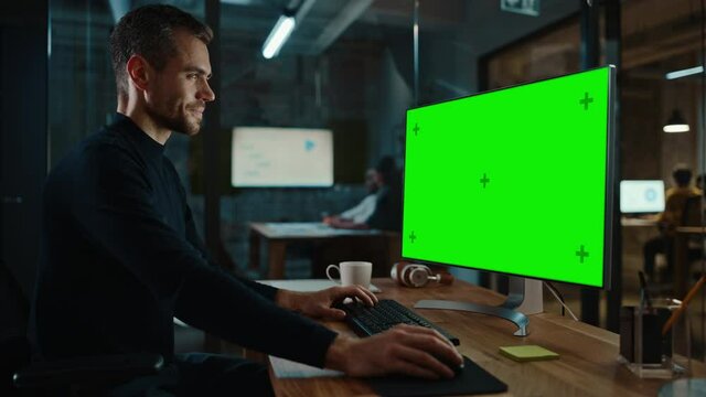 Young Handsome Specialist Working on Desktop Computer with Green Screen Mock Up Display in a Busy Creative Office with Colleagues. Male Manager with Trimmed Beard is Wearing a Casual Black Jumper.