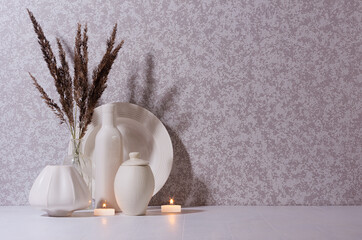 Evening cozy home decor with white ceramic vases, burning candles and bouquet of dried herb in moon light on white wood table, copy space.