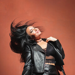Sexy brunette woman in leather jacket leather skirt and lace bralette posing red wall background hard light. Flying hair.