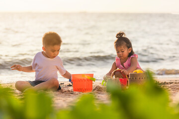 Happy fun two Asian children cute little boy and girl brother and sister family playing sand with toy sand tools at a tropical sea beach in holiday summer on sunset time, tourist trip concept