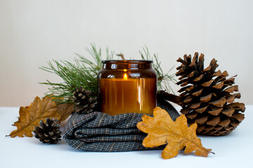 A large candle on a warm blanket, pine cones, autumn leaves on a white background. Cozy home. Autumn background. Autumn concept. Candle and decoration on a white background. Copy space. Yellow leaves