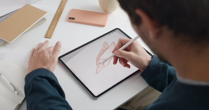 Over shoulder view of male artist drawing bird while creating illustration on note pad.Guy graphic illustrator using digital pad and stylus while sitting at table. Concept of modern art