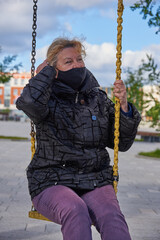 A 68-year-old woman wearing a black medical mask sits on a swing in the town square