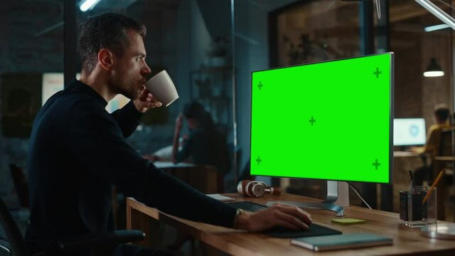 Young Handsome Specialist Working on Desktop Computer with Green Screen Mock Up Display in Creative Office. Male Manager Takes a Sip of Tea or Coffee from a White Mug He Picks Up from His Work Table.