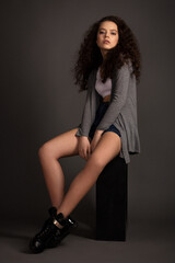 beautiful girl with skillful makeup and curly hair in a white sports bra and a gray cardigan sits on a black cube