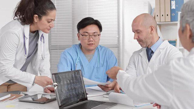 Over shoulder of unrecognizable grey-haired doctor looking at medical paper, passing in to young Asian male colleague across table. Multi Ethnic people talking, working in office