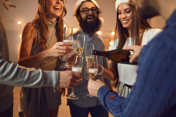 Group of young positive friends pouring champagne from bottle to glasses during Christmas party