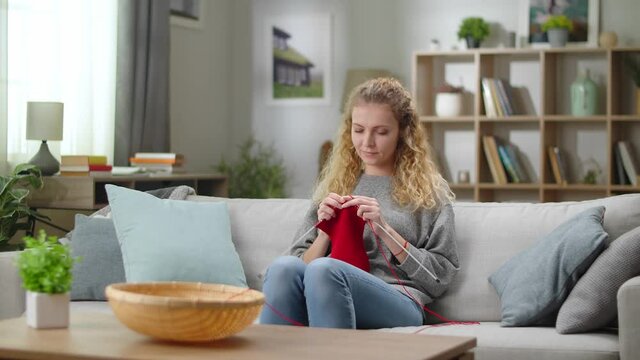 Woman sitting on sofa and use knitting needles in living room. Caucasian young woman knits socks made of red woolen threads. Quarantine hobby.
