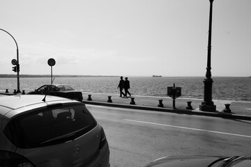 two older men go for a stroll down the pathway walkway by the water sea in black and white one is wearing a hat by the road with parked cars