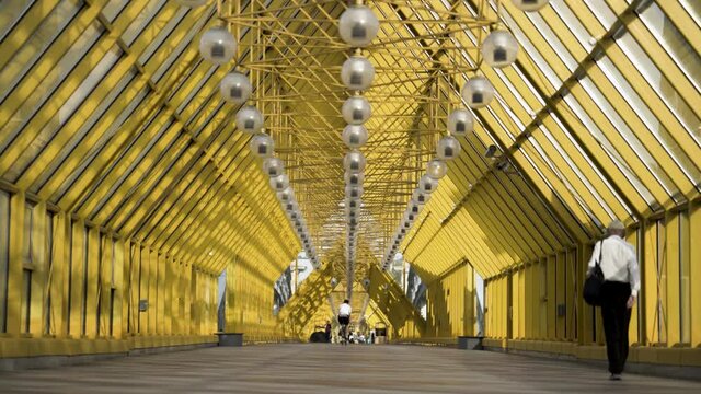 People walking inside pedestrian bridge in Moscow, Russia. Action. View inside of Andreyevskiy bridge with bright yellow metal beams and hanging lanterns.