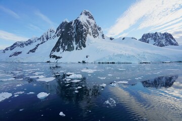 The rock is exposed at the top of the iceberg. The white clouds are radial. Some ice cubes are floating in the sea. Icebergs and white clouds reflected on the sea.