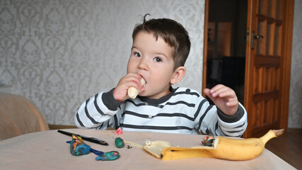 Naklejka premium Portrait of caucasian toddler boy at the table eating a banana. Snack during the game. Toddler boy spends time at home. 