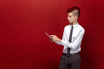 Portrait of red-haired teen punk with tablet against of red background. Isolated.