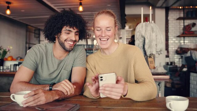 Young man laughing while sitting near girlfriend showing something funny on her smartphone