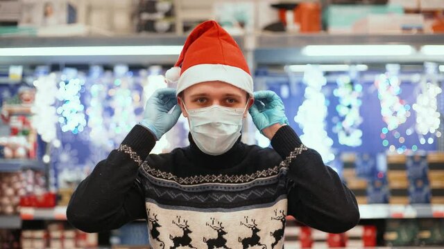 Young man in santa hat, medical gloves and christmas sweater puts on medical mask in supermarket. Shopping during pandemic covid-19 coronavirus concept