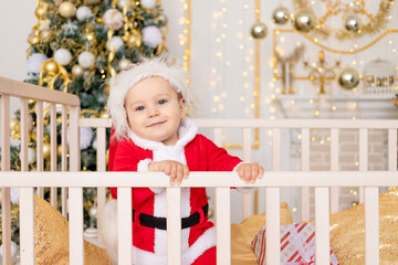 a child in a Santa costume stands in a crib at home near a Christmas tree with a Golden decor