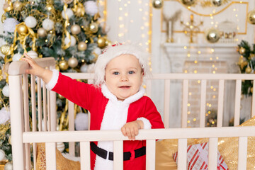 a child in a Santa costume stands in a crib at home near a Christmas tree with a Golden decor