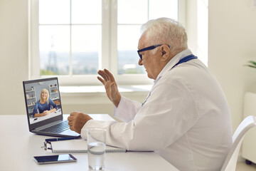 Fototapeta na wymiar Senior doctor sitting at desk, waving hand at laptop and saying hello to patient