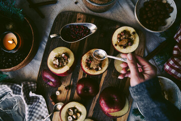 Process of making baked apples. Woman sprinkles brown sugar on fresh red apples stuffed with hazelnuts, raisins and lingonberries on wooden board on Christmas table.