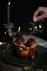 Pouring syrup on one of three baked apples stuffed with nuts, raisins and lingonberries on big green plate  on table decorated with candles, cones, fir branches on dark background .