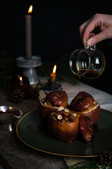 Pouring syrup on one of three baked apples stuffed with nuts, raisins and lingonberries on big green plate  on table decorated with candles, cones, fir branches on dark background .