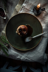 One baked apple stuffed with nuts, raisins and lingonberry, poured with syrup on big green plate with candle and saucer with fresh lingonberries and fir branches on wooden table.