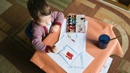 Little son painting cute home using colourful paints and sheet of white paper. Housing construction concept.