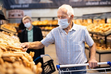 mature european man wearing mask and gloves with covid protection chooses buns and bread in supermarket bakery