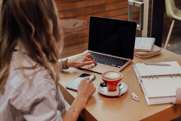 Fototapeta premium Woman working on a notebook in a cafe. Next to it is a cup of coffee and a planner. View from the back.