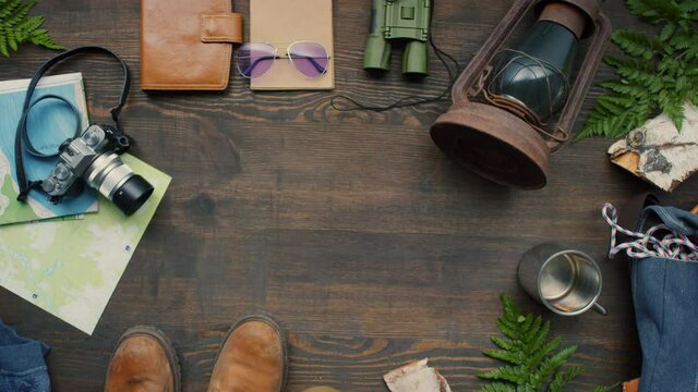 Top view flat lay footage of hiking boots, photo camera, rope, maps, backpack, lantern, notebooks, travel mug and binoculars lying on wooden table decorated with fern