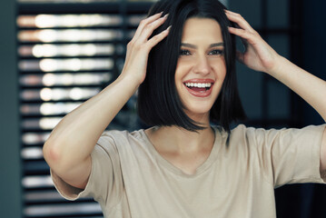 A positive brunette girl has a joyful expression during arranges her hair. Happy young woman looking to the mirror and making hairstyle.