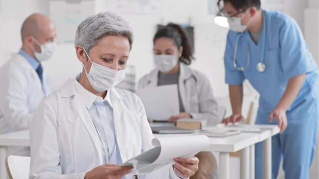 Waist up POV of senior silver-haired female Asian doctor in mask holding clipboard with papers, leafing through them, looking up on camera, blurred colleagues working around table on background